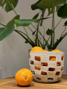 Panier Baguni en grès blanc fait main, rempli d'oranges, avec des motifs géométriques ajourés, posé sur une table en bois près d'une plante verte.
