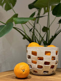 Panier Baguni en grès blanc fait main, rempli d'oranges, avec des motifs géométriques ajourés, posé sur une table en bois près d'une plante verte.