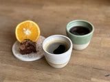Deux tasses expresso Kopi, une beige et une verte, remplies de café, accompagnées d'un gâteau et d'une tranche d'orange sur une table en bois.