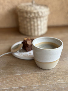 Tasse expresso Kopi en grès beige artisanal, posée sur une table en bois avec un gâteau et une tranche d'orange en arrière-plan.