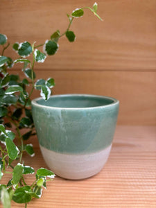 Tasse expresso Kopi verte en grès artisanal, posée sur une surface en bois avec une plante verte en arrière-plan.