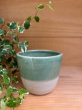 Tasse expresso Kopi verte en grès artisanal, posée sur une surface en bois avec une plante verte en arrière-plan.