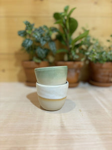 Deux tasses expresso Kopi empilées, une beige et une verte, sur une table en bois clair avec des plantes en arrière-plan.