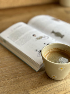 Tasse Expresso Marguerite avec motifs de fleurs blanches, posée sur une table en bois à côté d'un livre ouvert.