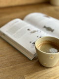 Tasse Expresso Marguerite avec motifs de fleurs blanches, posée sur une table en bois à côté d'un livre ouvert.