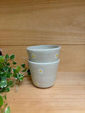 Deux Tasses Expresso Marguerite empilées, ornées de marguerites peintes, sur une surface en bois avec une plante verte en arrière-plan.