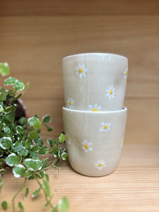 Deux Tasses Marguerite empilées, décorées de marguerites peintes à la main, posées sur une étagère en bois avec une plante verte.