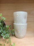 Deux Tasses Marguerite empilées, décorées de marguerites peintes à la main, posées sur une étagère en bois avec une plante verte.