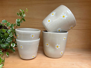 Ensemble de quatre Tasses Marguerite artisanales en grès blanc, décorées de marguerites, disposées sur une étagère en bois avec une plante.