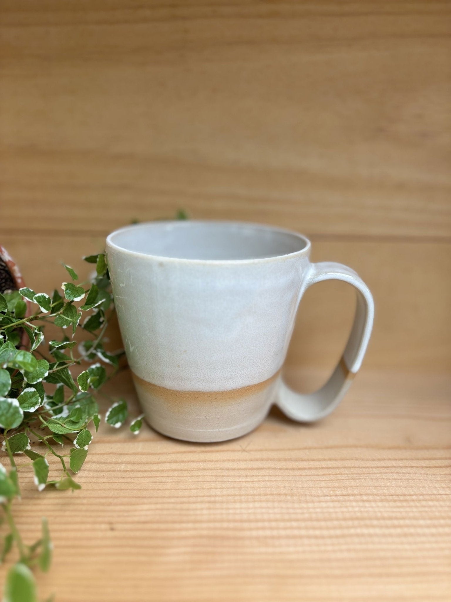 Tasse Nubi au design minimaliste en grès blanc avec une finition artisanale, placée sur une surface en bois avec des feuilles vertes en arrière-plan.