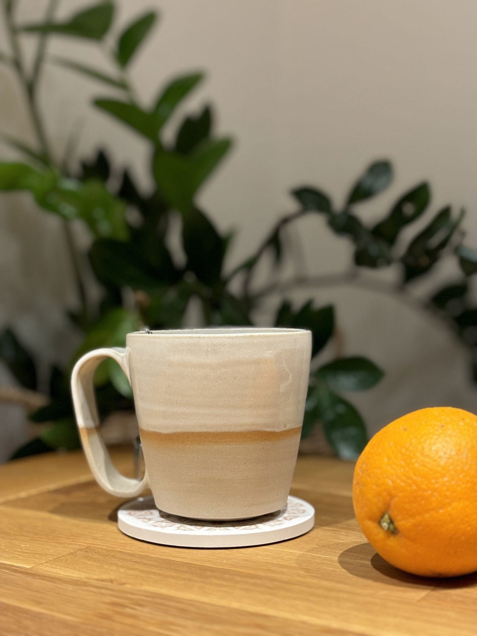 Tasse Nubi en grès blanc de 35cl avec design épuré artisanal, posée sur une table en bois près d'une orange et d'une plante verte.
