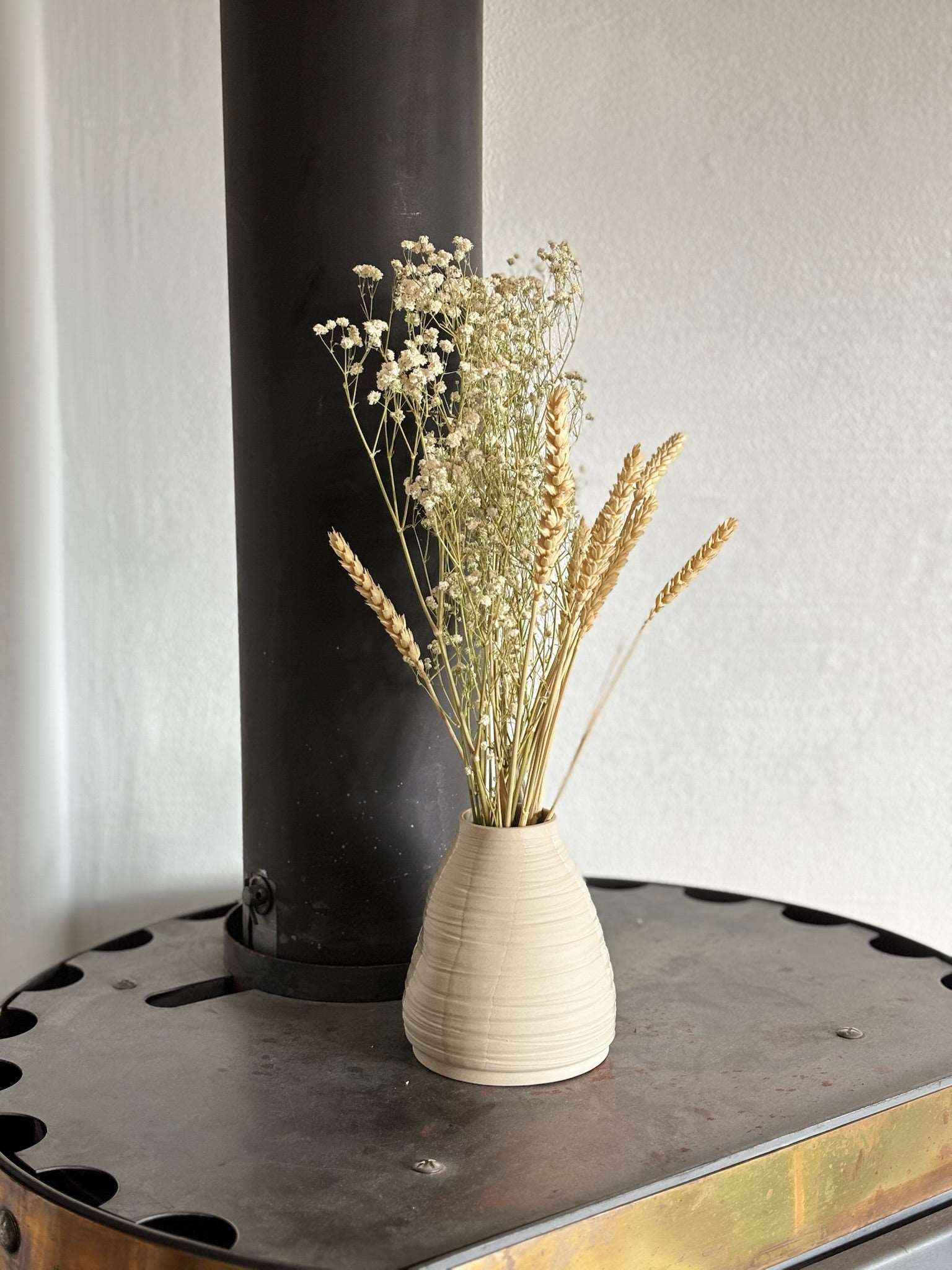 Vase Pado artisanal en céramique beige avec des fleurs séchées, posé sur un poêle noir. Idéal pour une décoration minimaliste.